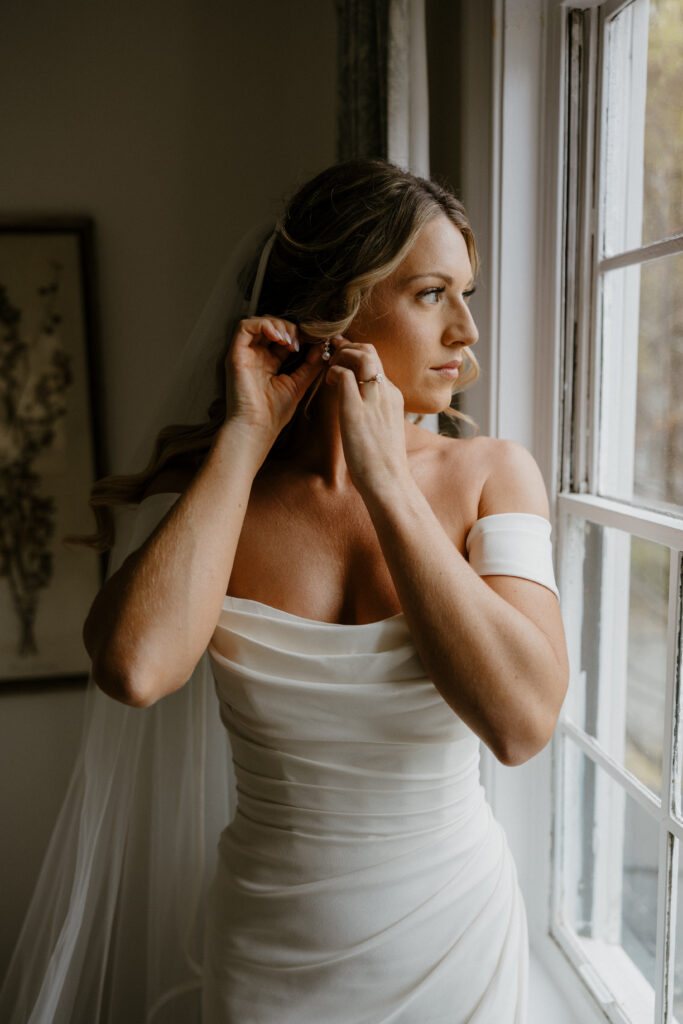 bridal getting ready photo of her putting on her earring with her wedding dress on looking out the window