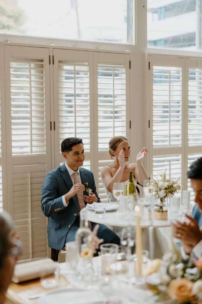 bride and groom sitting at their sweetheart table together clapping and laughing together 