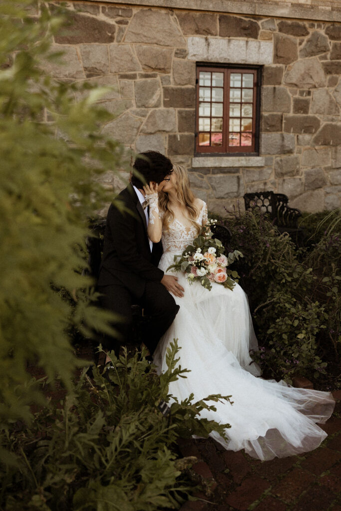 bride and groom sitting down together kissing while bride is holding wedding bouquet 